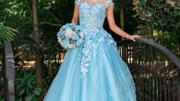 A young woman standing gracefully in a blue ball gown with a tiara surrounded by lush greenery.