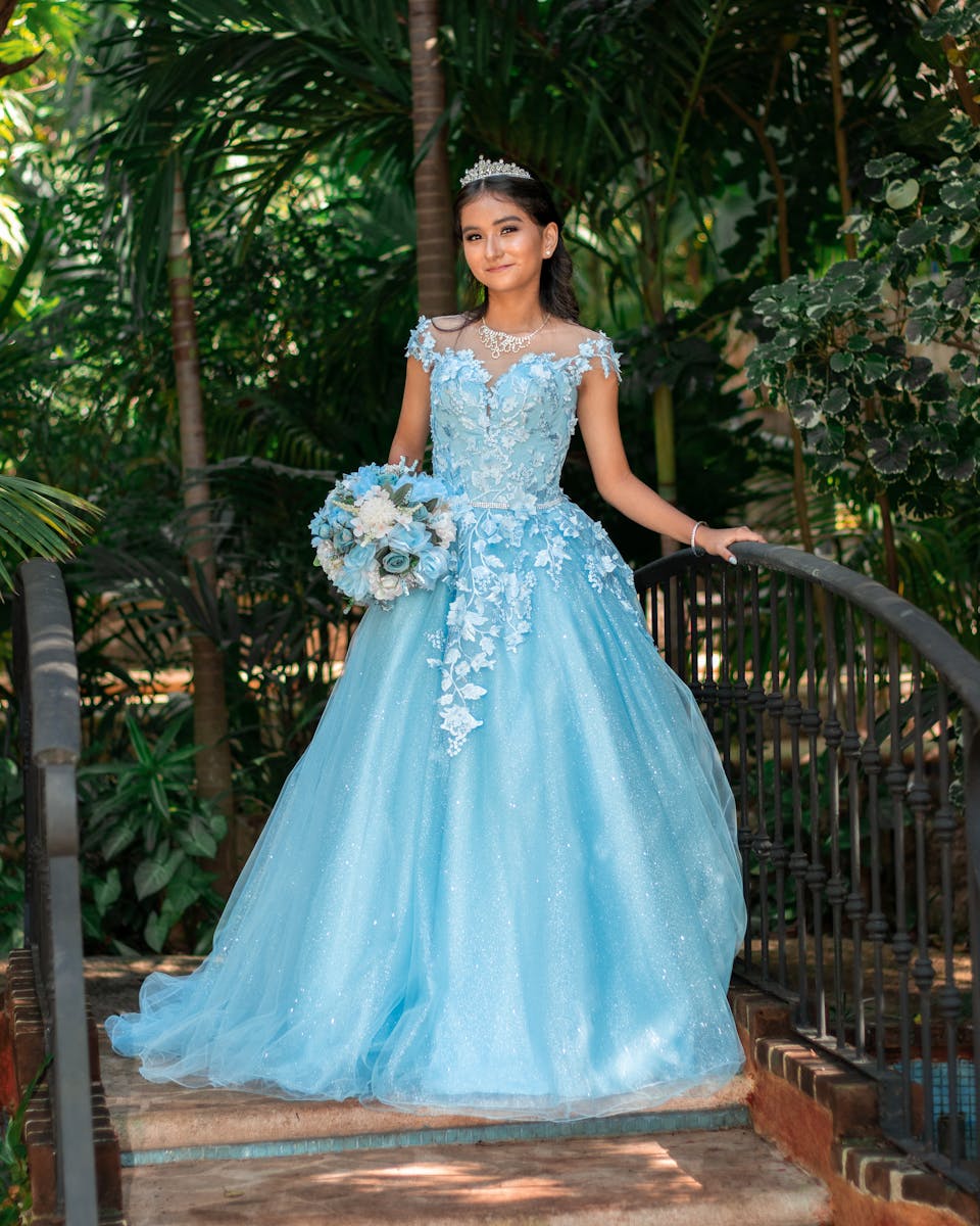 A young woman standing gracefully in a blue ball gown with a tiara surrounded by lush greenery.