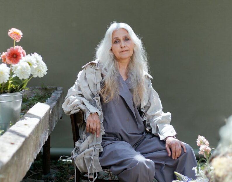 Senior woman in gray sitting indoors surrounded by flowers, creating a serene scene.