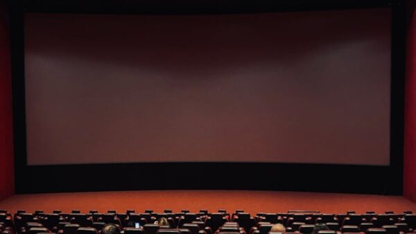 Dimly lit cinema hall with red seats and a blank screen. Minimal audience.
