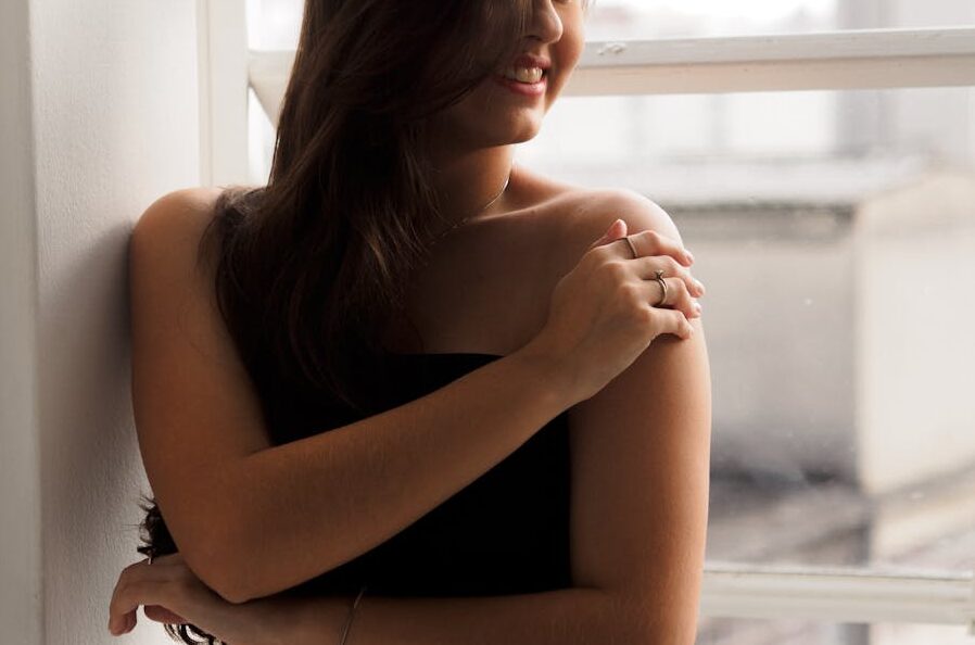Elegant woman in a black dress standing by a window, smiling softly.