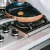 Crop faceless woman breaking raw egg into frying pan on gas stove while preparing breakfast in light kitchen at home