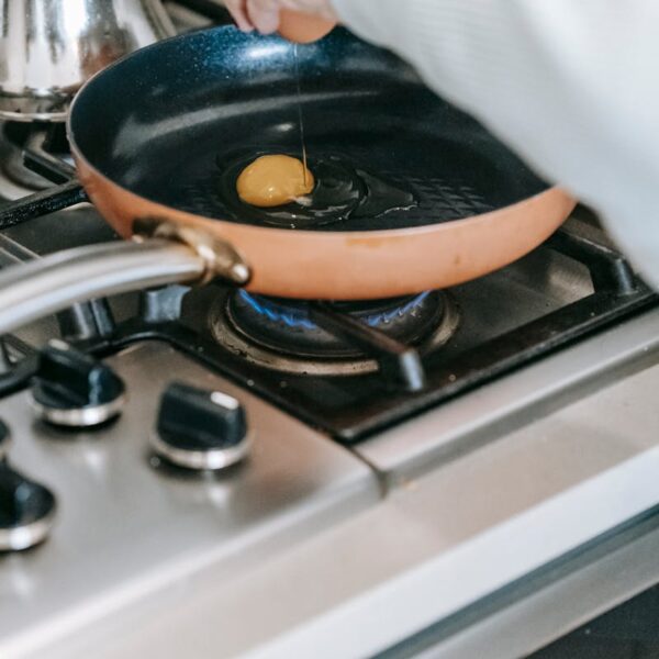 Crop faceless woman breaking raw egg into frying pan on gas stove while preparing breakfast in light kitchen at home