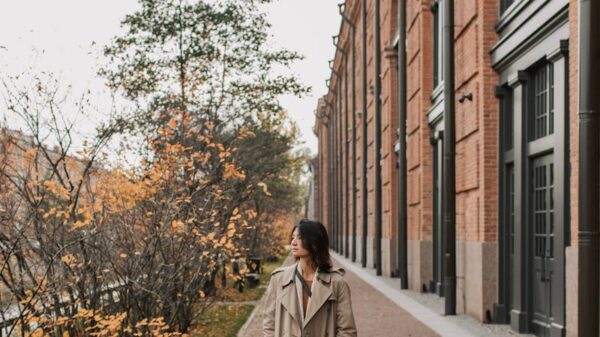 Woman in a trench coat walking along a tree-lined city street amidst autumn foliage.