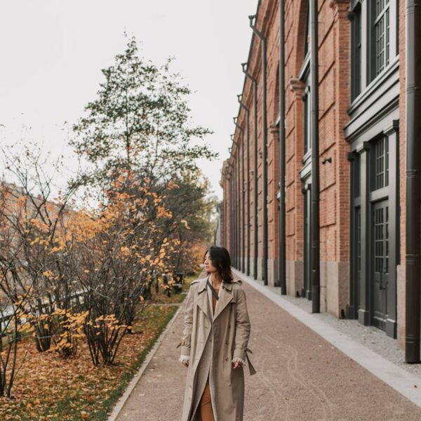 Woman in a trench coat walking along a tree-lined city street amidst autumn foliage.