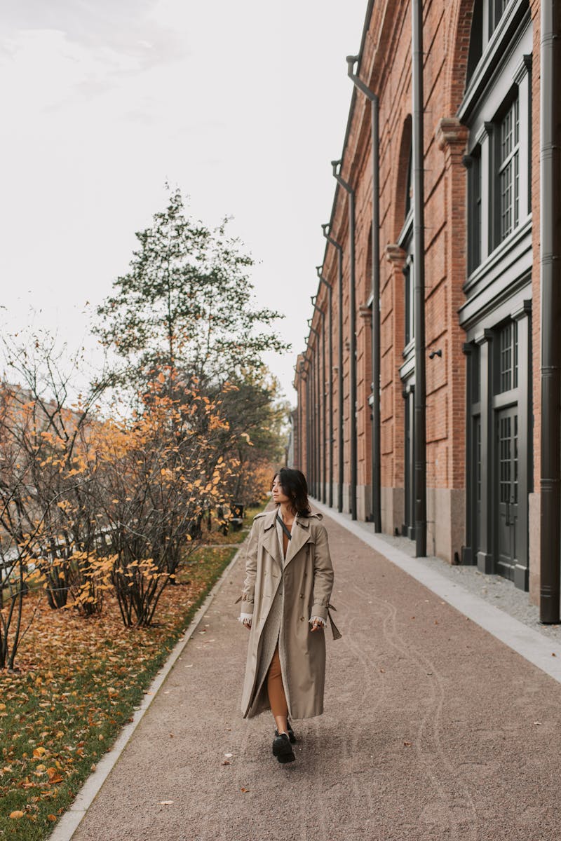 Woman in a trench coat walking along a tree-lined city street amidst autumn foliage.