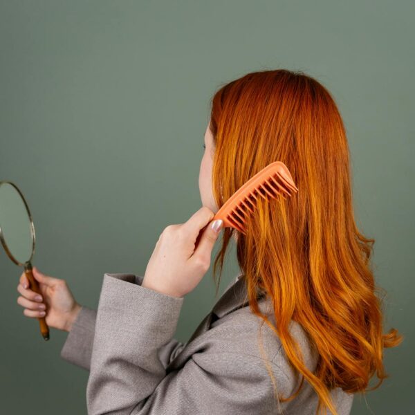 Red-haired woman using a comb while holding a mirror, focusing on hair care and beauty.