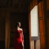 A woman in a red gown poses gracefully near an ornate window, exuding elegance.