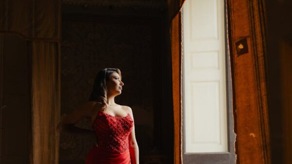 A woman in a red gown poses gracefully near an ornate window, exuding elegance.