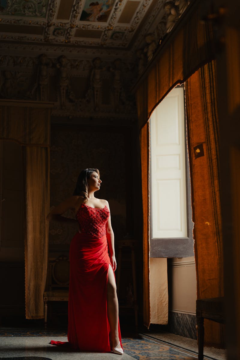 A woman in a red gown poses gracefully near an ornate window, exuding elegance.
