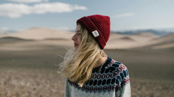 Side view of young female in casual warm sweater and red knitted hat standing in dry desert