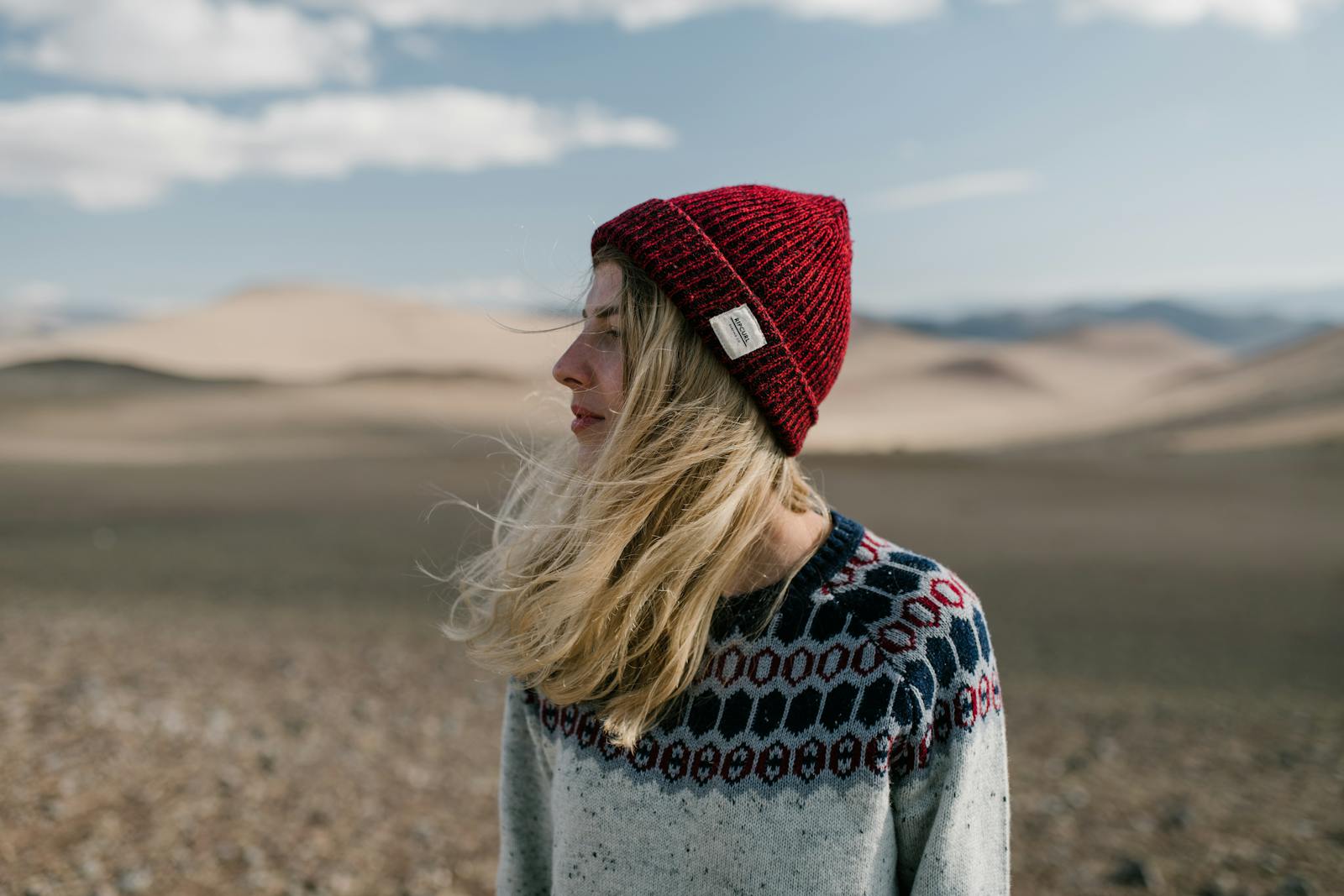 Side view of young female in casual warm sweater and red knitted hat standing in dry desert