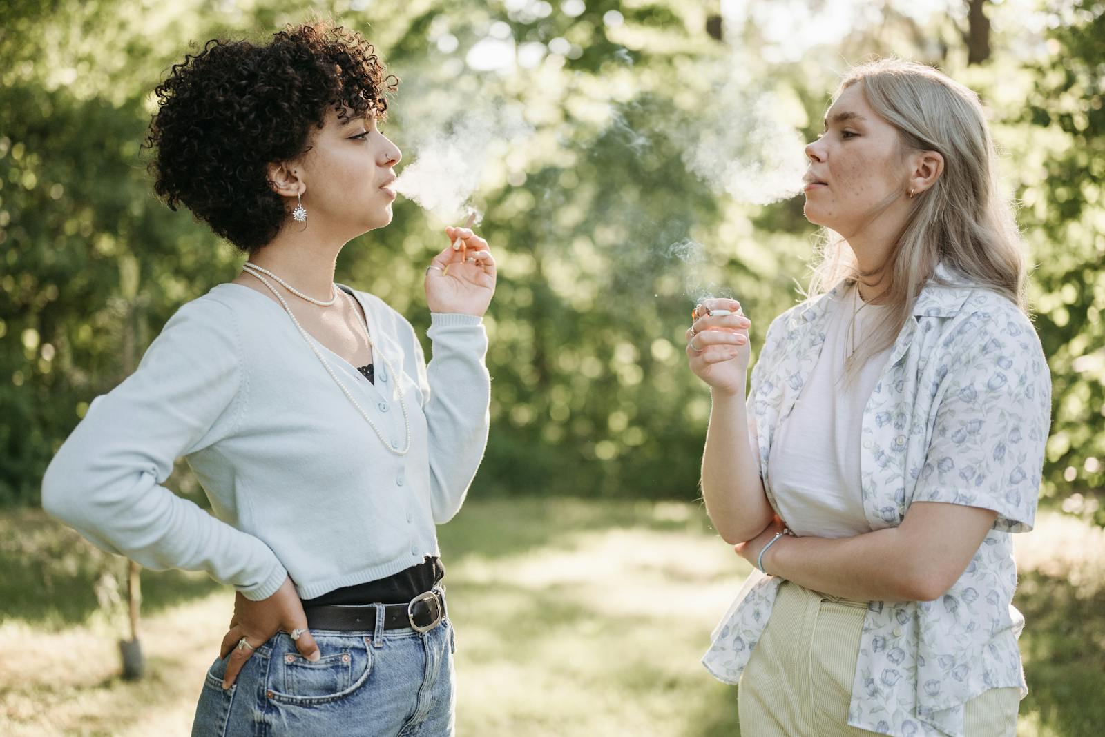 Two women smoking and conversing in a lush green outdoor setting, enjoying a moment of relaxation.