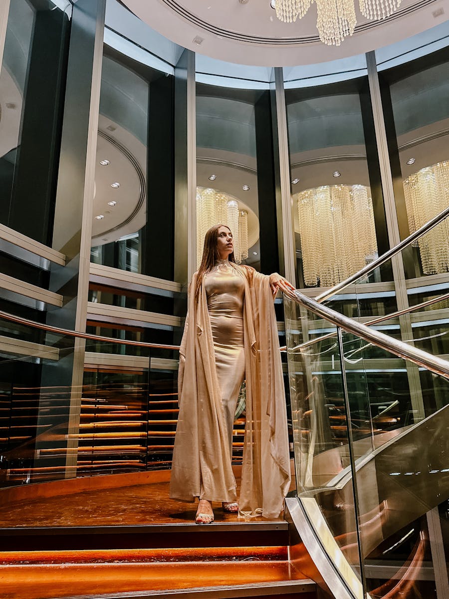 Graceful woman in a satin dress poses on a luxurious indoor staircase, surrounded by modern architecture.