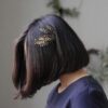 Profile view of a woman with a bob haircut adorned with a gold hairpin, against a neutral background.