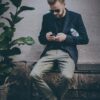 man using smartphone sitting on black metal bench near brown concrete wall