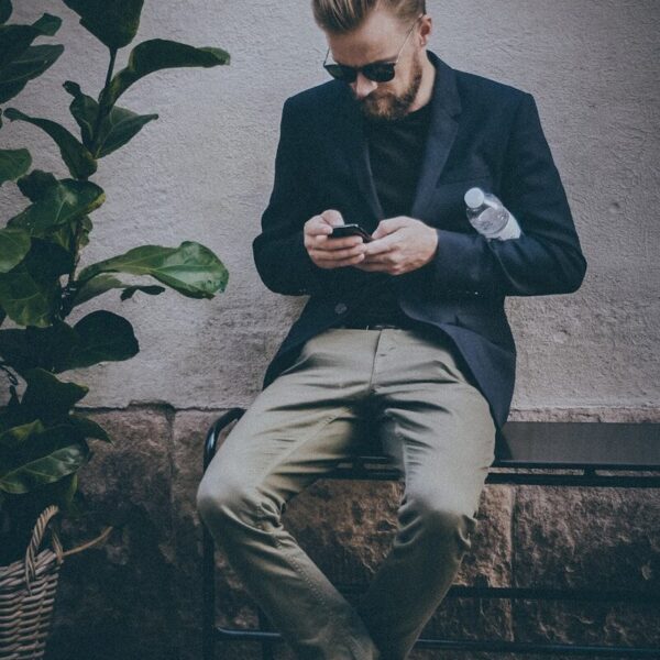 man using smartphone sitting on black metal bench near brown concrete wall