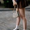 A young woman wearing a fashionable outfit holds a chic plaid bag, standing on an urban street.
