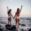 two women standing on rock while raising their hands near sea during daytime