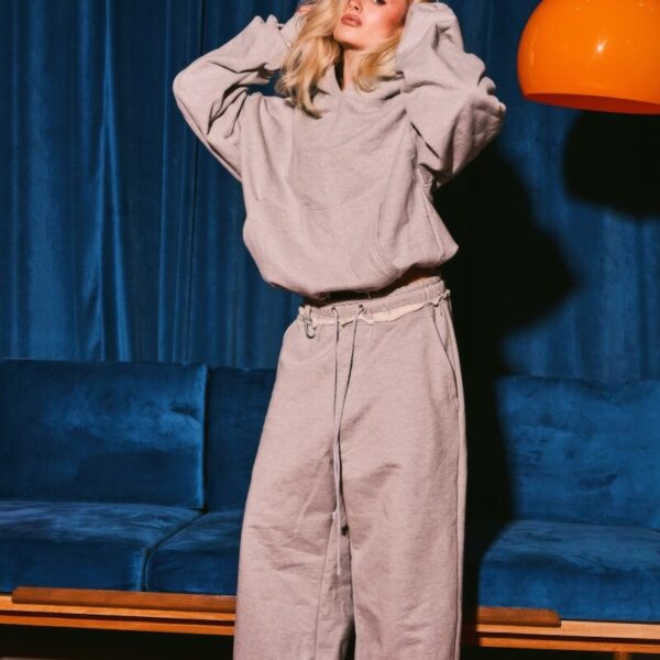Woman in grey hoodie and pants posing against blue background.