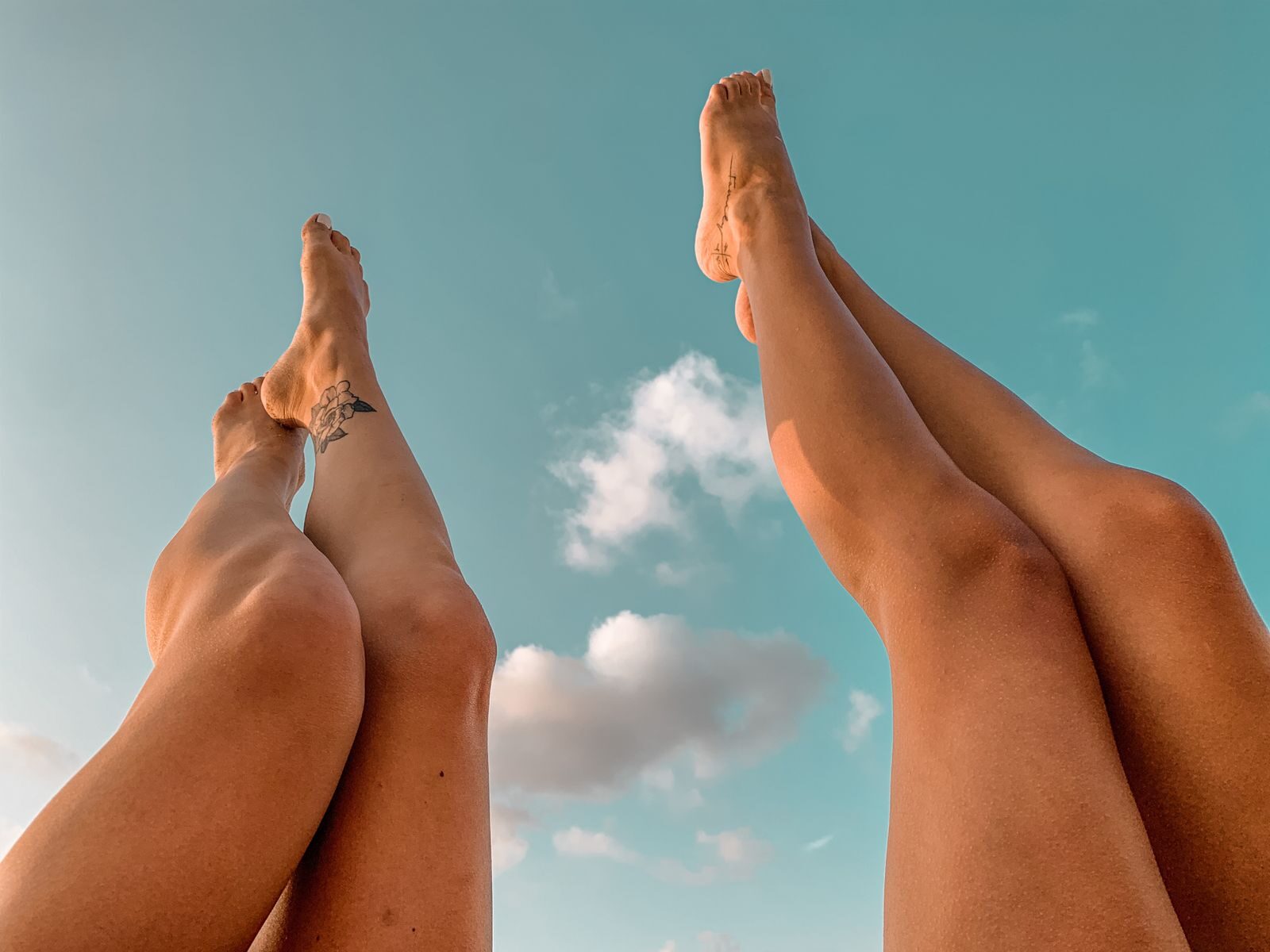 Artistic photo of two women's legs against a clear blue sky with clouds, emphasizing freedom and summer vibes.